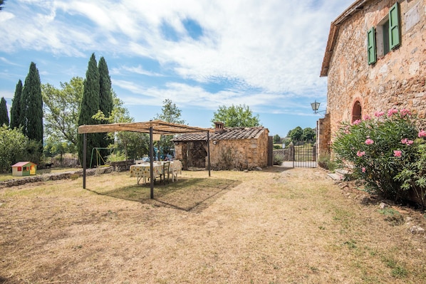A rustic stone building with a thatched roof garden area, a small wooden structure, and a paved path.