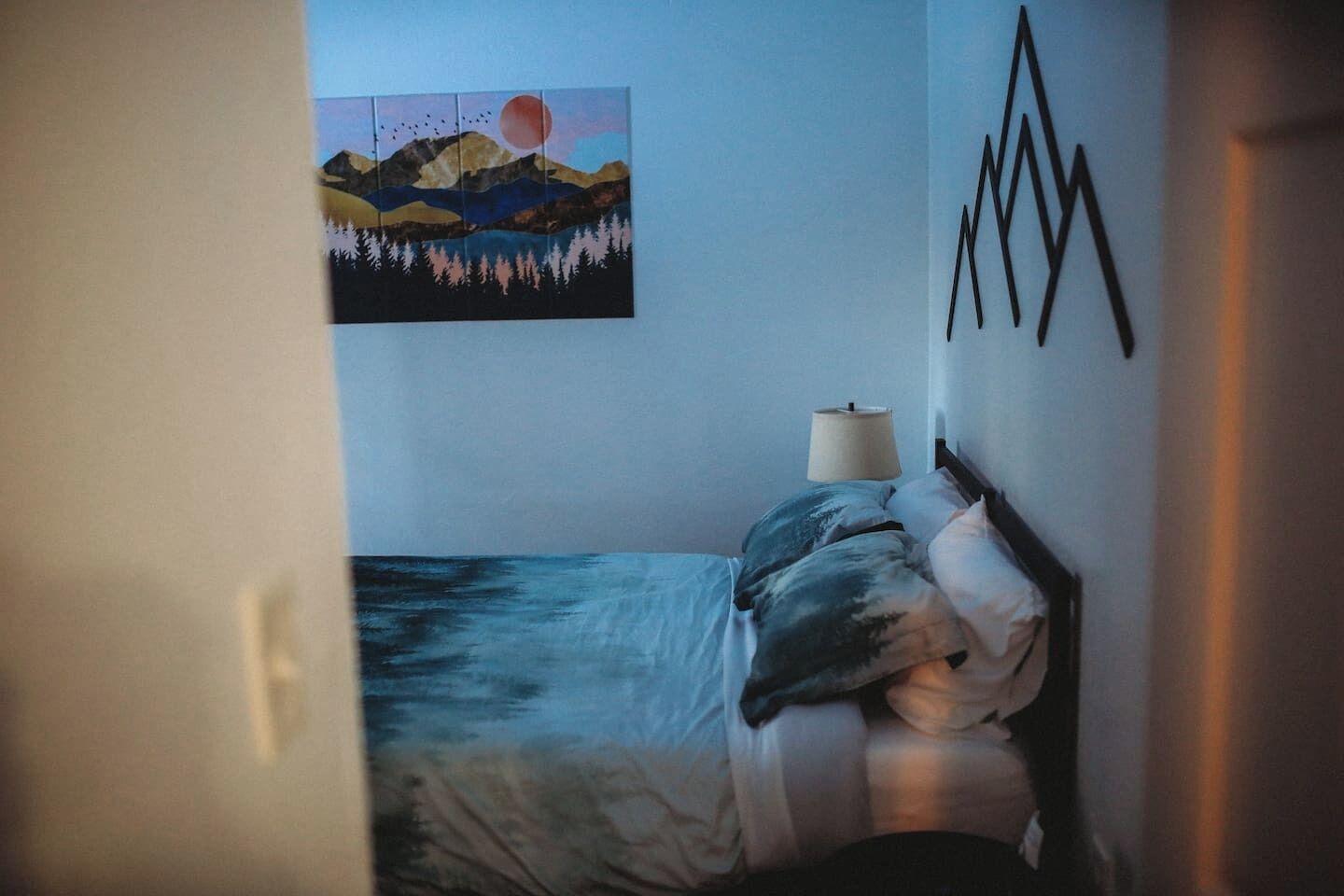 Un dormitorio con cama, mesita de noche, lámpara y un cuadro de paisaje en la pared.
