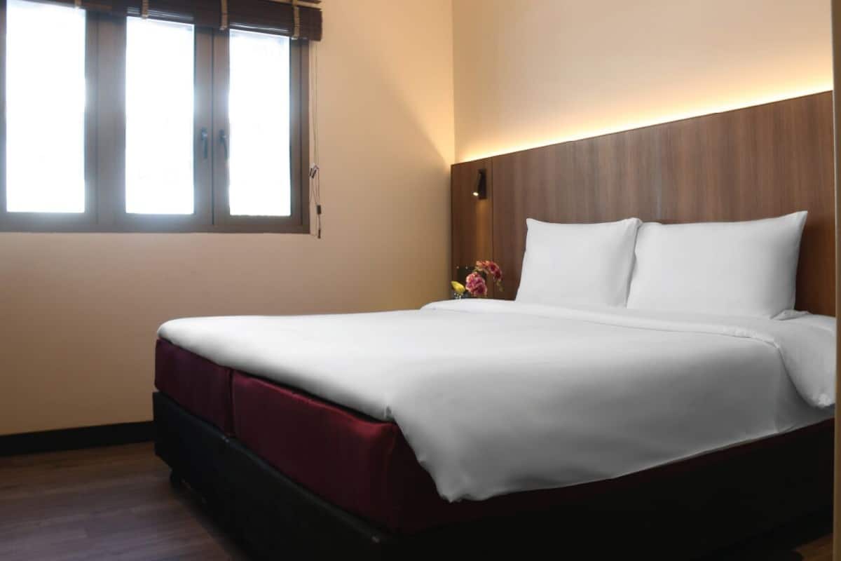 Une chambre d’hôtel avec un lit, une fenêtre et une table de chevet ornée d’un vase de fleurs.