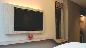 Une chambre d’hôtel dotée d’un téléviseur à écran plat fixé au mur, d’un miroir au-dessus et d’un petit bol de pommes posé sur une étagère en dessous.
