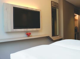 Habitación de hotel con televisión de pantalla plana montada en la pared, un espejo encima y un frutero pequeño con manzanas en un estante debajo.