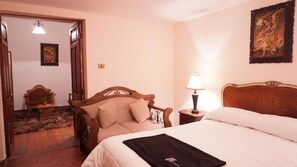 A bedroom with a bed, a wooden chair, a nightstand with a lamp, and a painting on the wall. - Museo Hacienda Cayara (POTOSI)
