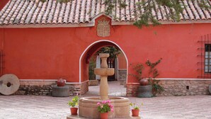 Un patio con una fuente de piedra, plantas en macetas y un edificio de techos de tejas y paredes rojas.