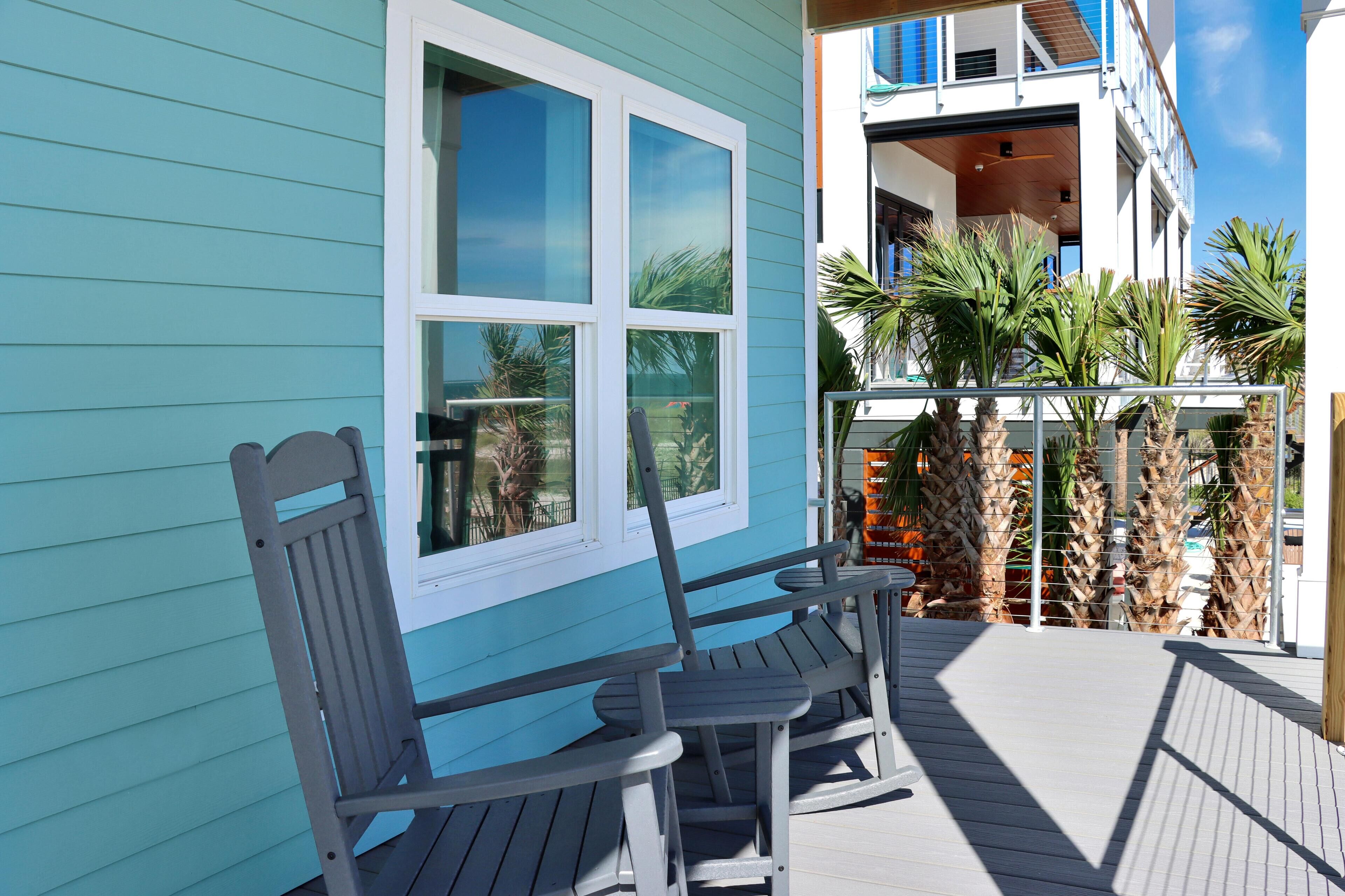 A wooden deck with two rocking chairs and a window with a view of palm trees.