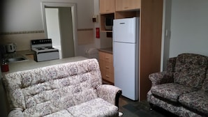 Un espace de vie compact comprenant un canapé, un fauteuil, un réfrigérateur, une cuisinière et un petit coin cuisine.