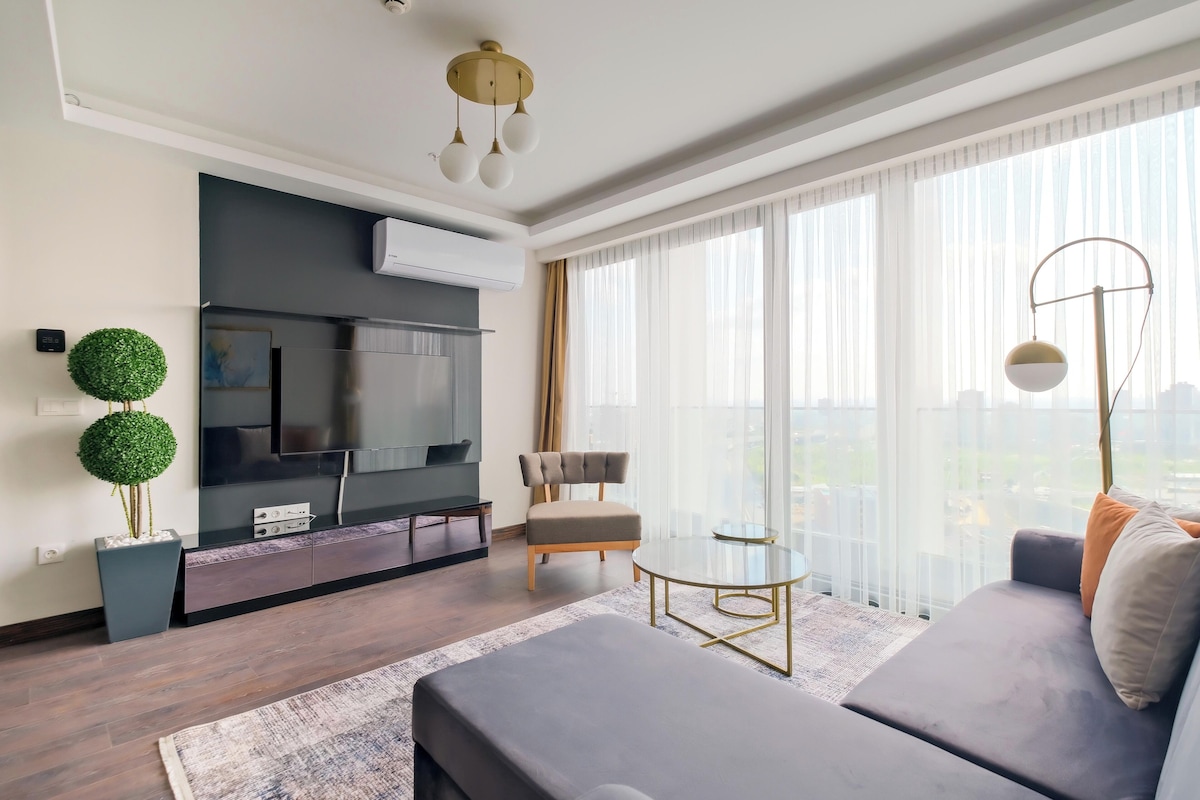 A modern living room with a flat-screen TV, a sofa, a round coffee table, and a large window with sheer curtains.