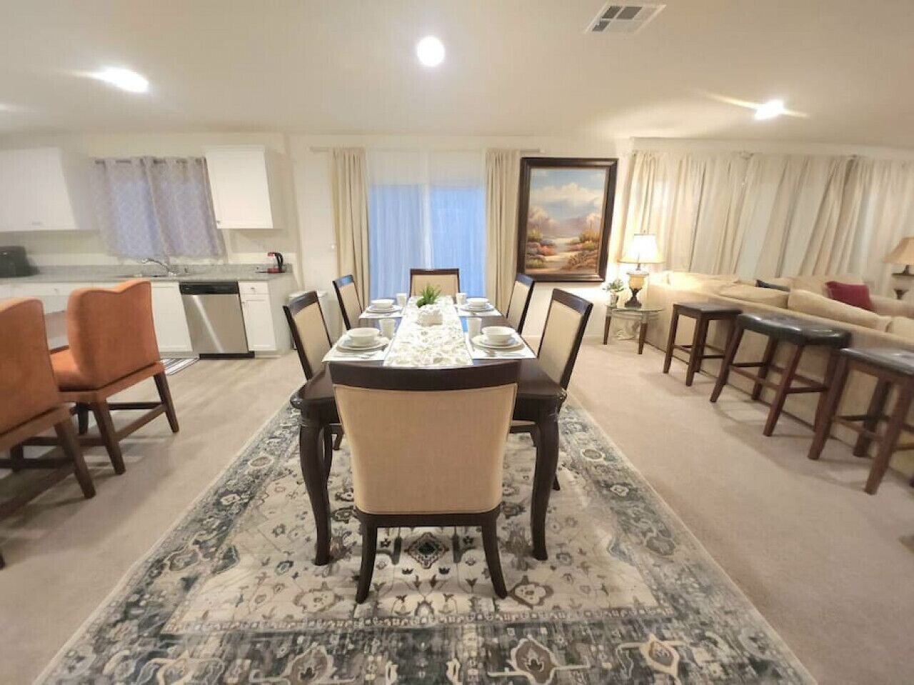 A dining area with a table set for six, surrounded by chairs, a kitchen in the background, and a sofa with a lamp.