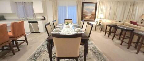 A dining area with a table set for six, surrounded by chairs, a kitchen in the background, and a sofa with a lamp.