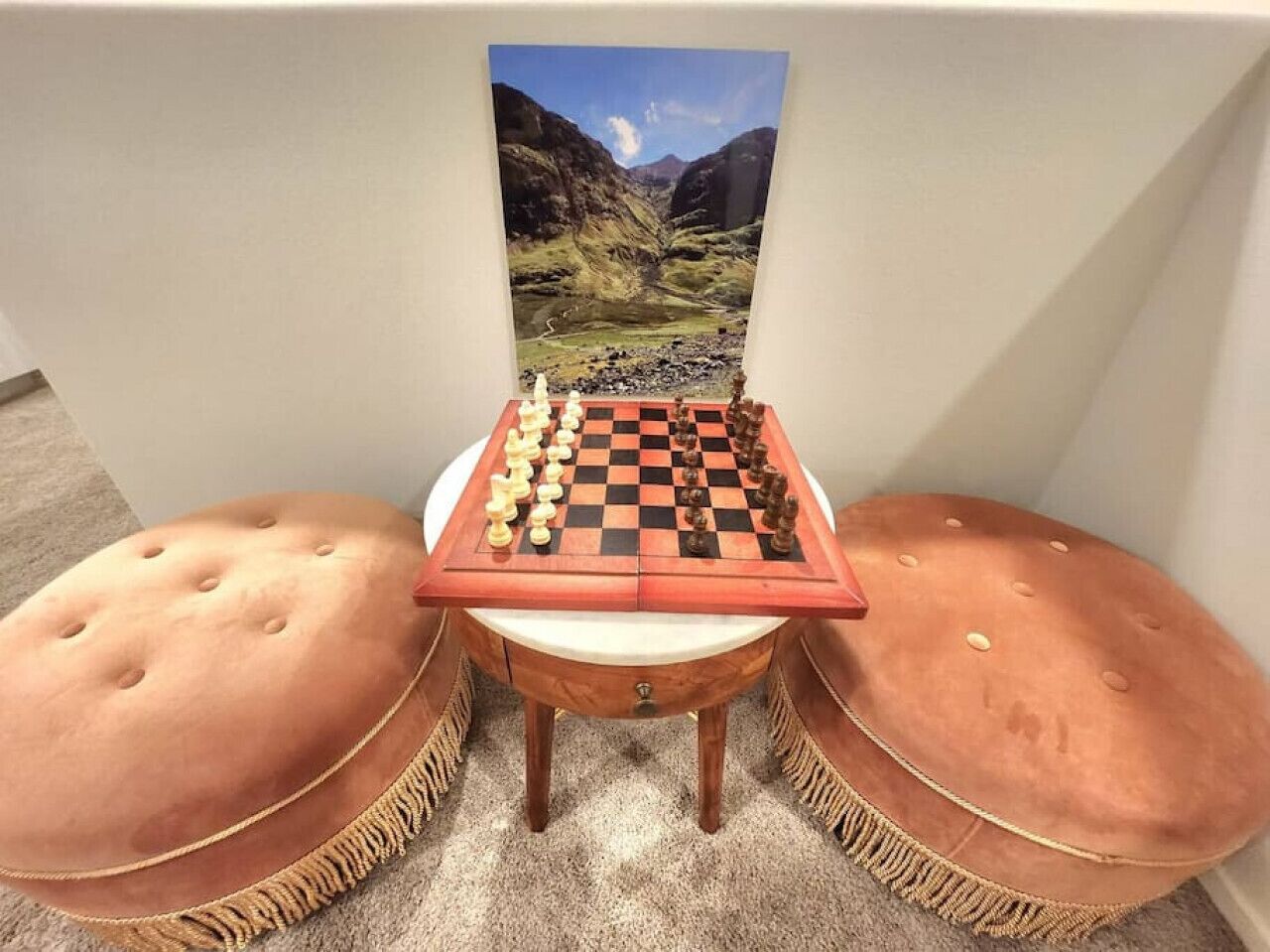 A chessboard set on a small table with a scenic landscape poster behind it, flanked by two cushioned stools.