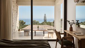 A bedroom with a large sliding glass door leading to a balcony with a view of the sea and a tree.