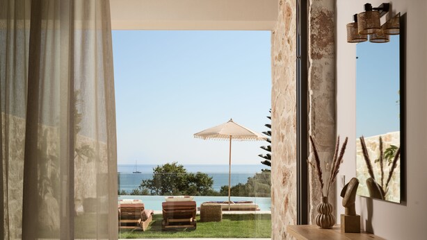 A modern room with a large window overlooking a pool and ocean view. There is a wooden bench with cushions and a large umbrella.