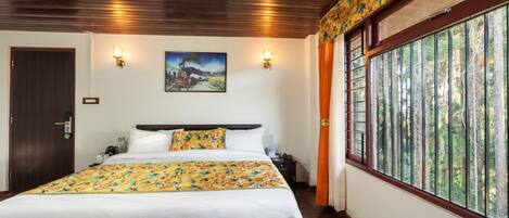 Quarto com cama grande, teto de madeira, duas luzes fixadas na parede, um quadro e janela com vista para árvores.