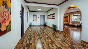 Un hall spacieux doté d’un parquet en bois, une grande peinture aux motifs floraux sur le mur de gauche, et un coin salon avec un canapé et une fenêtre en arrière-plan.