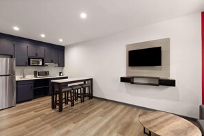 A modern kitchen with dark cabinets, a microwave, a refrigerator, and a dining area with a wooden table and stools. - Ramada By Wyndham Studio Suites Dothan (Dothan)