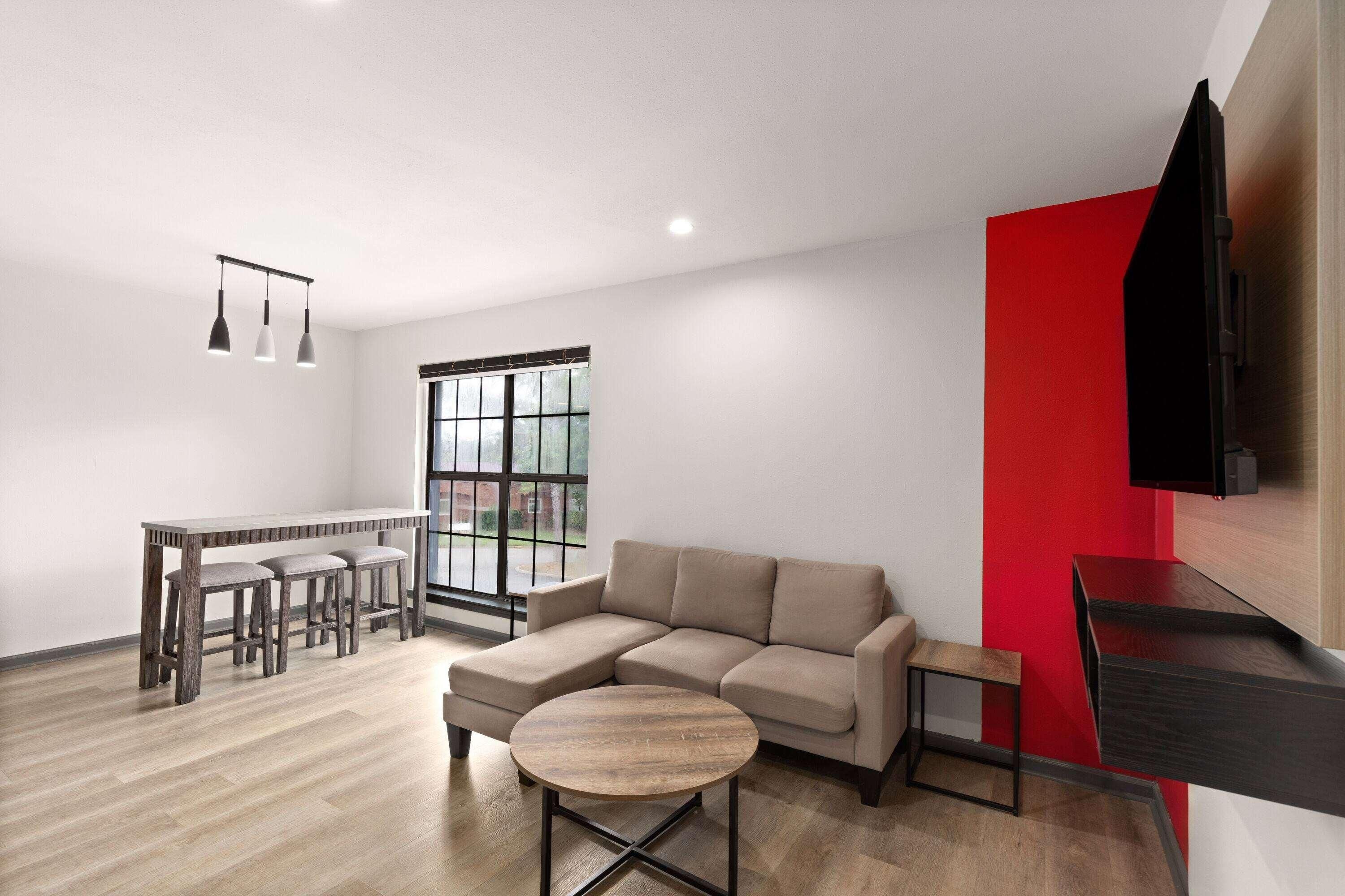 A modern living room with a sectional sofa, a round coffee table, a dining area with a bench, and a TV mounted on a red accent wall.