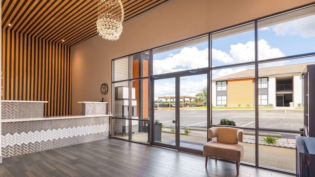 A reception area with a wooden reception desk, a large window with a view of a parking lot and buildings, and a hanging light fixture. - Ramada By Wyndham Studio Suites Dothan (Dothan)
