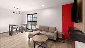 A modern living room with a sectional sofa, a round coffee table, a dining area with a bench, and a TV mounted on a red accent wall. - Ramada By Wyndham Studio Suites Dothan (Dothan)
