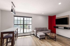 A modern living room with a sofa, a coffee table, a TV mounted on the wall, and a dining area with a table and chairs. - Ramada By Wyndham Studio Suites Dothan (Dothan)