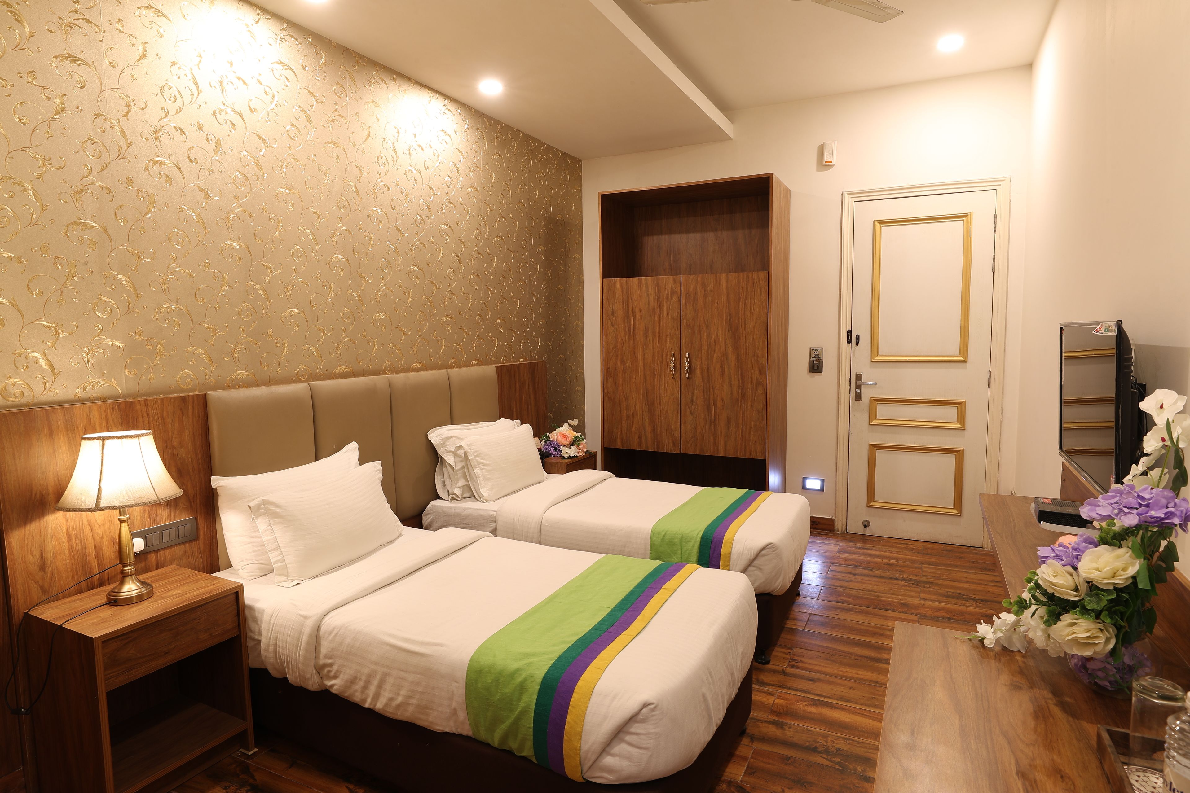 A hotel room with two beds, a nightstand with a lamp, a wooden wardrobe, and a desk with a vase of flowers.