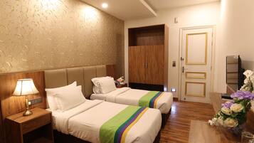 A hotel room with two beds, a nightstand with a lamp, a wooden wardrobe, and a desk with a vase of flowers.