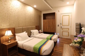 A hotel room with two beds, a nightstand with a lamp, a wooden wardrobe, and a desk with a vase of flowers. - Antilia By Zion (Sonepat)