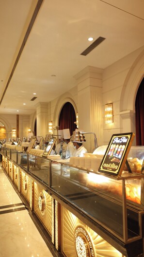 A luxurious hotel lobby with a long counter displaying various food items, elegant chandeliers, and a grand arched doorway. - Antilia By Zion (Sonepat)