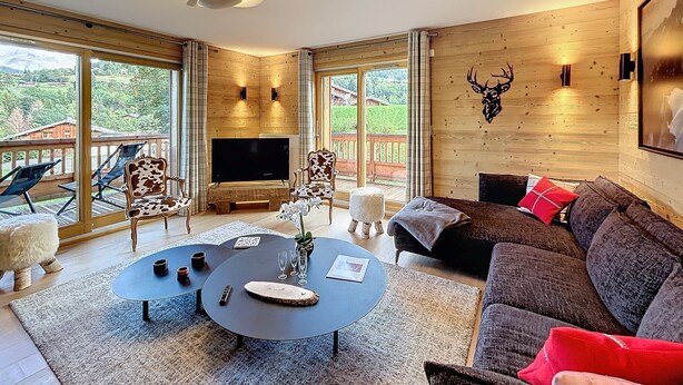 Un salón amplio con televisor grande, paredes de madera, cabeza de venado montada y balcón con mobiliario exterior.