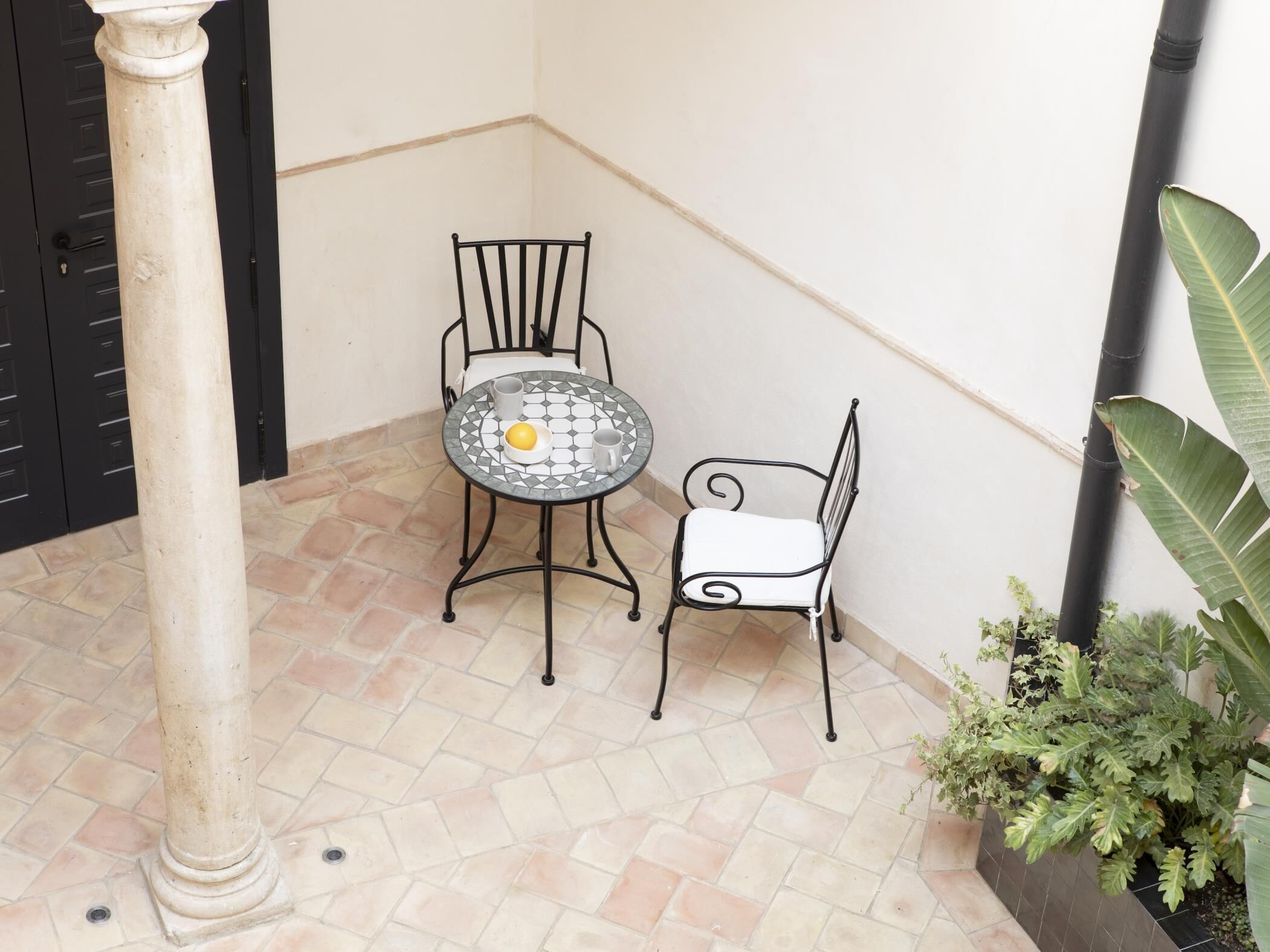A small patio area with a tiled floor, a round table with a yellow object on it, two metal chairs, and a potted plant.