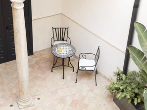 A small patio area with a tiled floor, a round table with a yellow object on it, two metal chairs, and a potted plant.