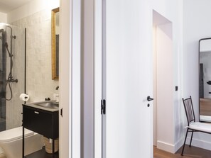 Ein modernes Badezimmer mit Duschkabine aus Glas, einer weißen Toilette, einem schwarzen Waschbecken mit Spiegel darüber und einem weißen Stuhl im angrenzenden Raum.