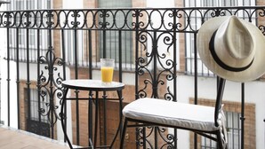 Een balkon met een sierijzeren hekwerk, een kleine ronde tafel met een glas sinaasappelsap, een witte fauteuil met kussens en een hoed die aan het hekwerk hangt.