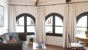 Ein modernes Wohnzimmer mit einer Couch, einem Couchtisch mit einem Laptop, einem Esstisch mit Stühlen und großen Bogenfenstern mit Vorhängen.