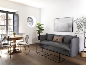 A modern living room with a grey sofa, a round coffee table, and a dining table with chairs. There is a large window with a view of buildings, a potted plant, and a framed artwork on the wall.
