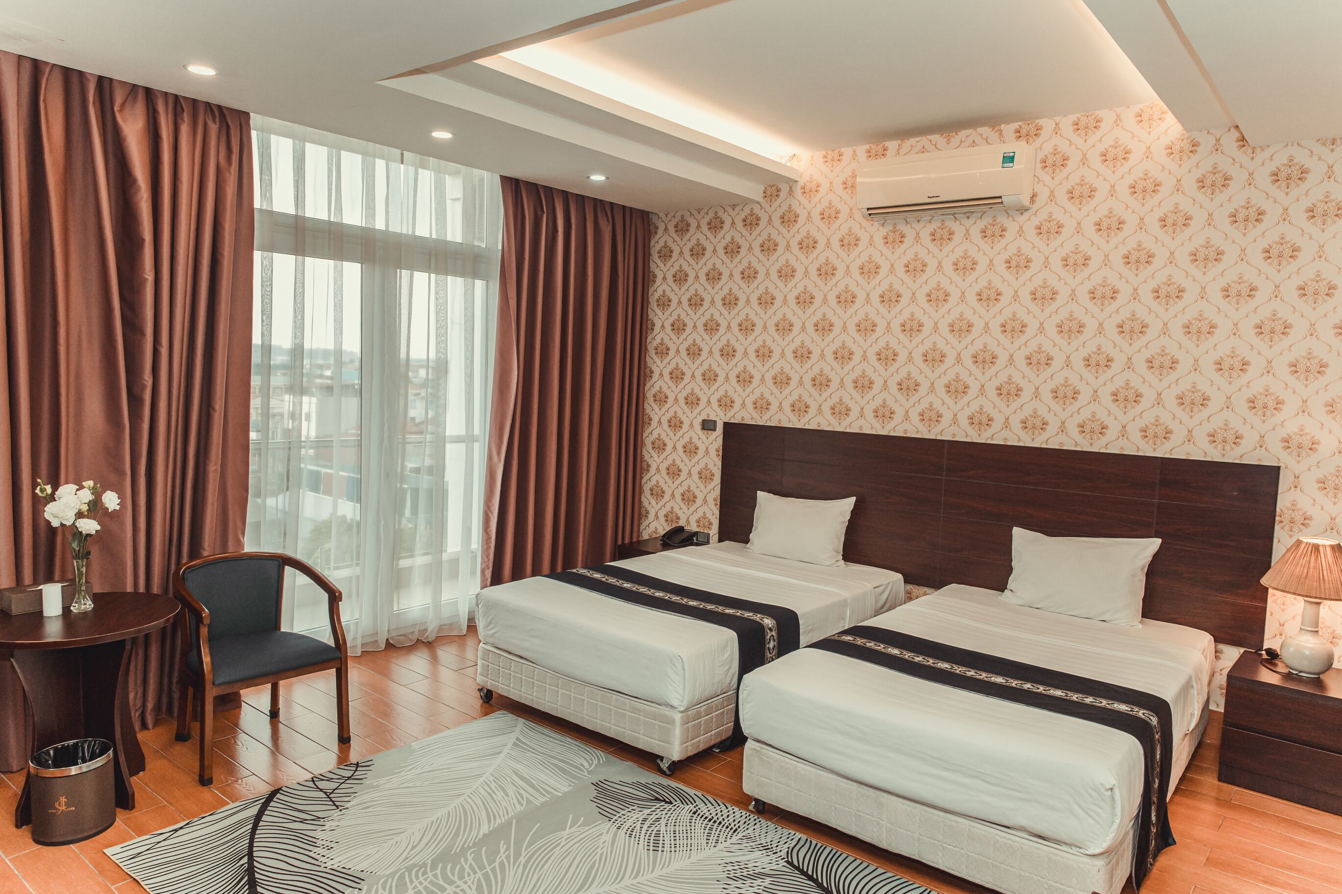 Habitación de hotel con dos camas, un escritorio con silla, una mesita con un jarrón de flores y una alfombra estampada.