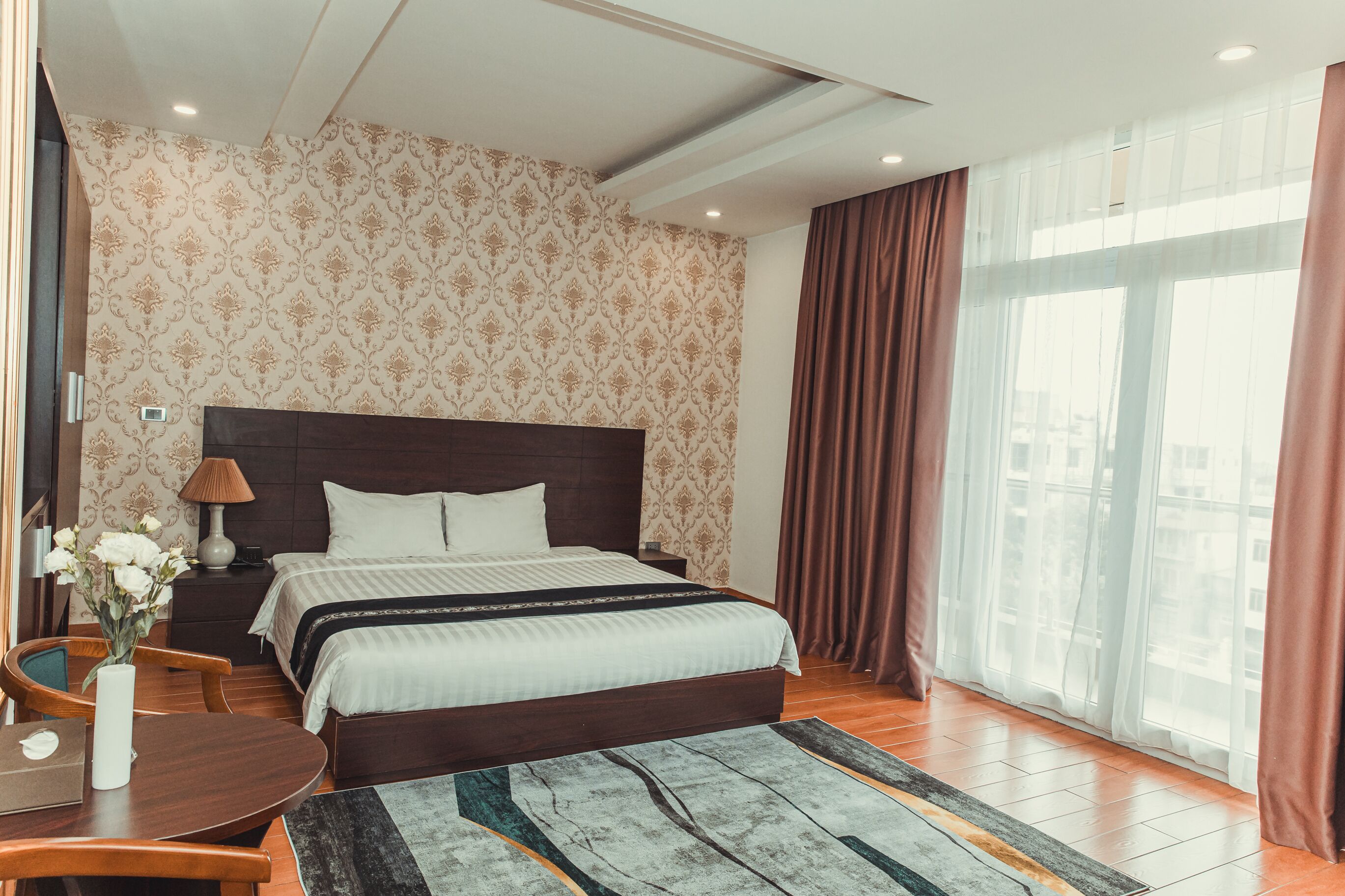 A hotel room with a large bed, a desk with a chair, a small table with a vase of flowers, and a window with curtains.
