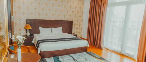Habitación de hotel con una cama grande, cabecero de madera, mesita de noche con lámpara, una pequeña mesa con un florero y vistas a la ciudad a través de amplias ventanas.