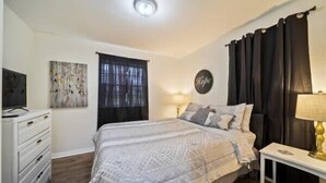 A bedroom with a bed, a dresser, a TV, a painting, a window with curtains, and a wall-mounted lamp. - Spacious 4/2BR Home |Pet-Friendly |Midtown Tulsa (Tulsa)