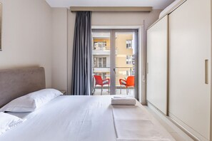 A hotel room with a bed, a window with curtains, a balcony with chairs, and a sliding door.