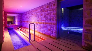 Un espace piscine intérieure aux murs en pierre, doté d’une terrasse en bois et d’une rambarde métallique.