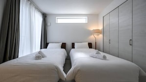 Quarto de hotel com duas camas, um guarda-roupa espaçoso, uma janela com cortinas e uma luminária fixada na parede.