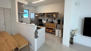 Uma cozinha moderna com eletrodomésticos embutidos, um espaço de refeições com mesa e banco de madeira, e uma sala de estar com televisão e plantas decorativas.