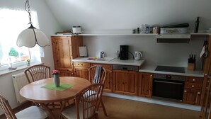 A kitchen with wooden cabinets, a dining table with chairs, a hanging light fixture, and a window with a view of greenery outside. - Vacation apartment in the countryside between Binz and Sellin (Zirkow)