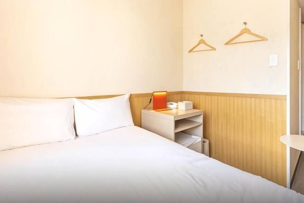 A hotel room with a bed, a bedside table with a lamp, a small shelf, and two wooden hangers on the wall.
