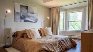 Un dormitorio con una cama grande, una mesita de noche, una lámpara, una ventana con cortinas y un cuadro en la pared.