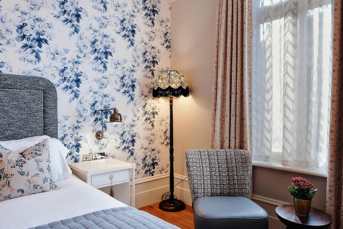 Quarto com papel de parede floral, mesa de cabeceira, luminária, cadeira estampada e janela com cortinas.