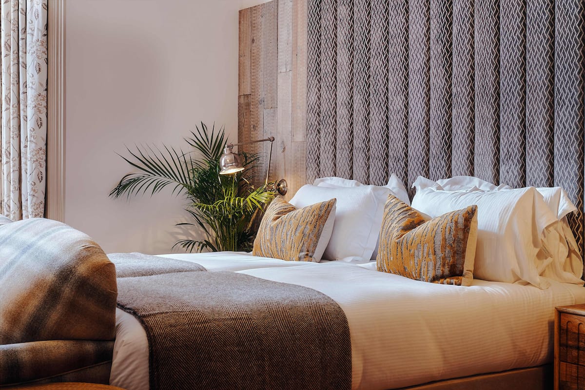 Cama bem arrumada com lençóis brancos e almofadas estampadas, um vaso de planta e um abajur em uma mesa de cabeceira.