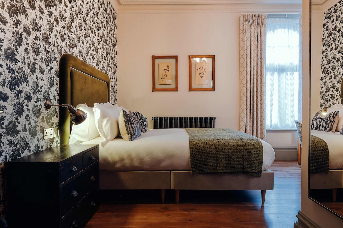 Quarto com cama, cómoda, duas gravuras emolduradas de plantas e janela com cortinas.