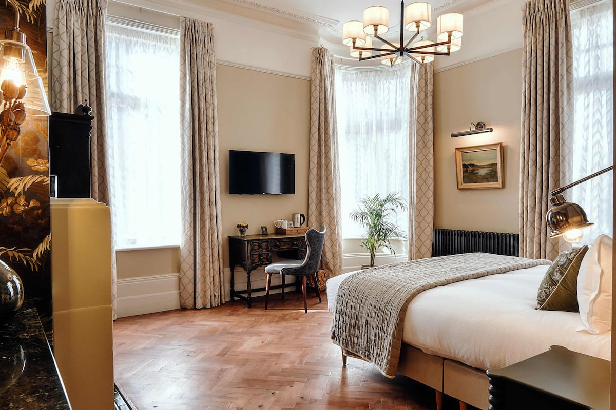 Quarto de hotel com cama, escrivaninha, cadeira, televisão e uma grande janela com cortinas.
