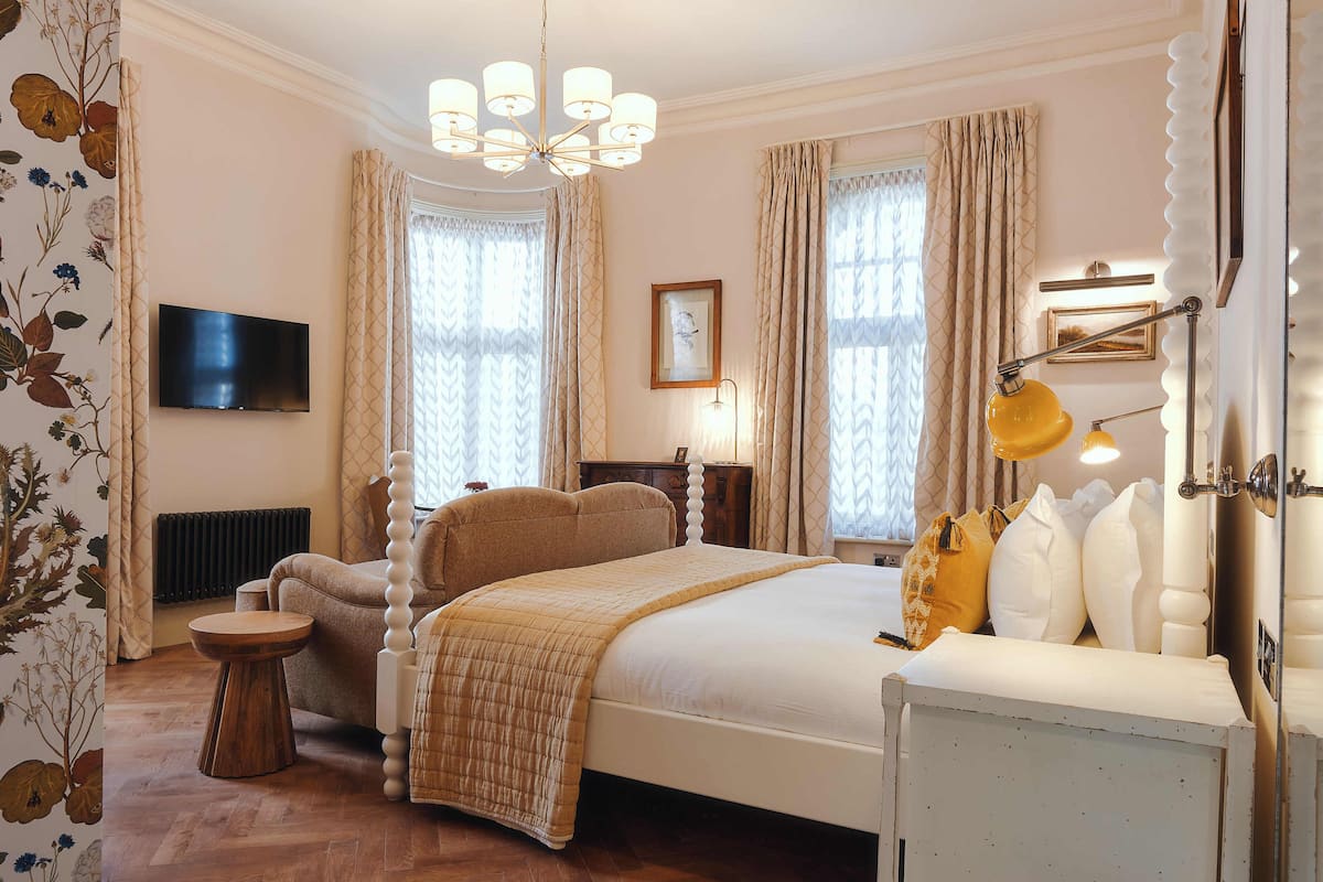 Quarto com cama, mesa de cabeceira, abajur, televisão e cortinas.