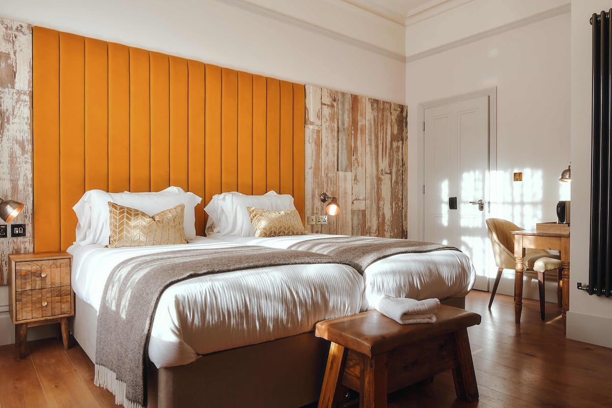 Quarto com cama grande, cabeceira de madeira, mesas de cabeceira e um banquinho de madeira.